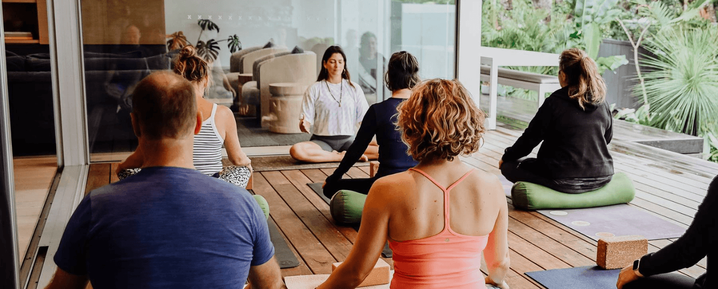 Yoga teacher trainees sitting cross legged in meditation on a sunny timber deck