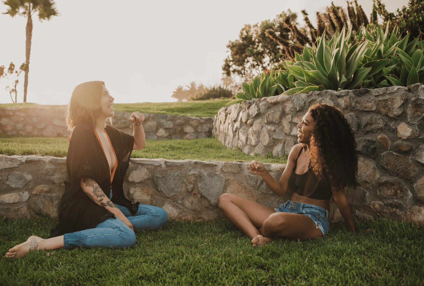 Two women laughing together on the grass beside a stone wall in warm afternoon light